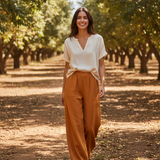 Woman standing in an orchard wearing a white top and brown pants.