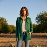 Woman wearing a green jacket standing in an outdoor setting with trees and clear sky.