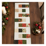 A Christmas-themed quilted table runner with a patchwork design of red, green, beige, and black patterned fabric squares, displayed on a dark wooden dining table surrounded by holiday decorations.
