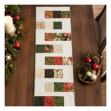 A Christmas-themed quilted table runner with a patchwork design of red, green, beige, and black patterned fabric squares, displayed on a dark wooden dining table surrounded by holiday decorations.