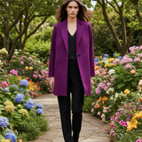 A woman walks down a stone garden path wearing an oversized purple blazer over a black top and black trousers, surrounded by colorful flowers and green trees.