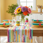 A colorful watercolor-style table runner draped over a wooden dining table, styled with yellow plates, teal napkins, glassware, and a vibrant flower bouquet in a bright kitchen setting.