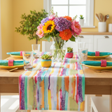 A colorful watercolor-style table runner draped over a wooden dining table, styled with yellow plates, teal napkins, glassware, and a vibrant flower bouquet in a bright kitchen setting.